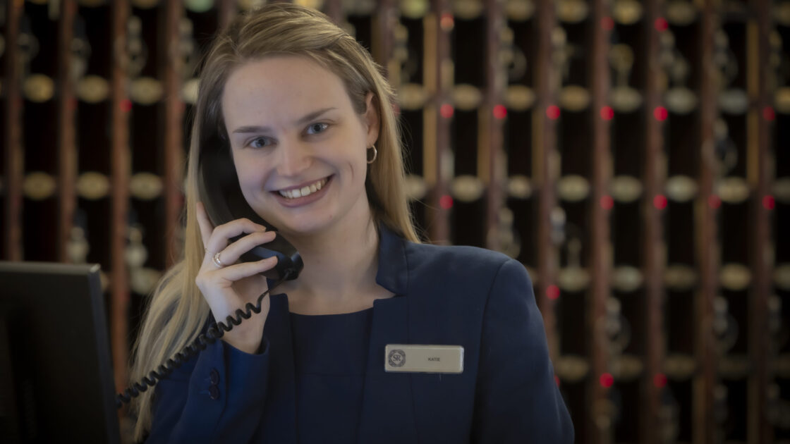Friendly hotel receptionist on phone with a welcoming smile at the elegant front desk, ready to assist guests.