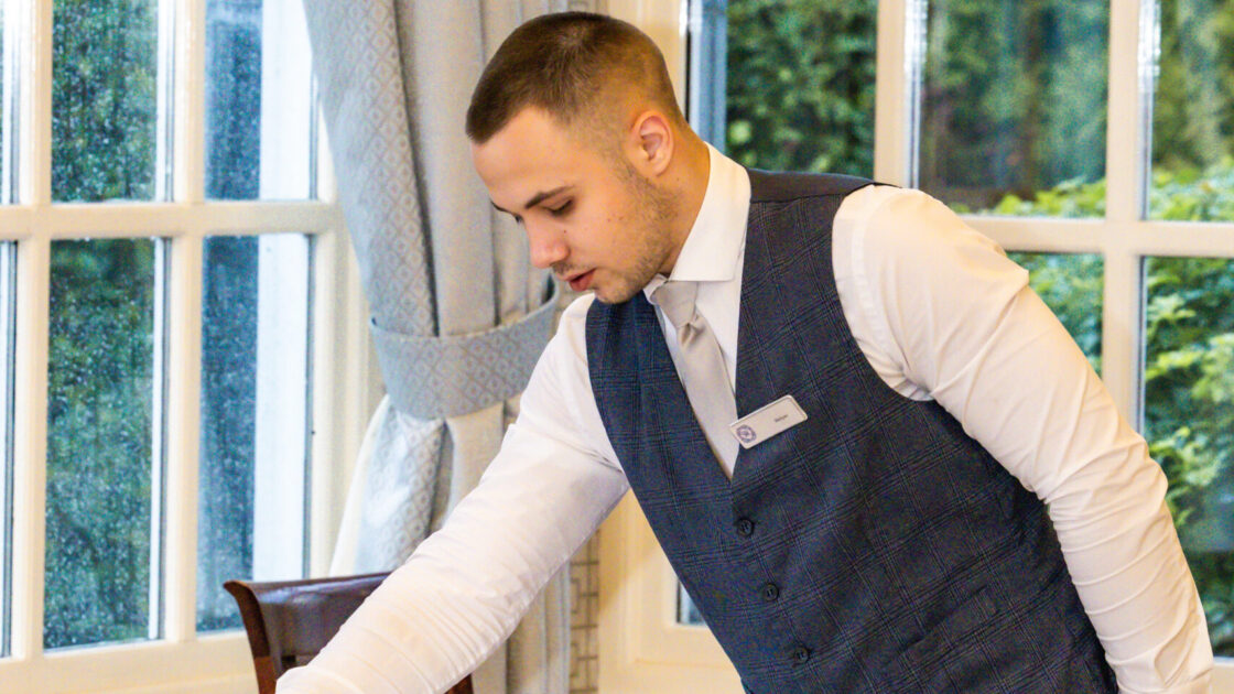 Waiter setting an elegant dining table by large windows in a bright hotel restaurant, ready for guests to enjoy fine dining.