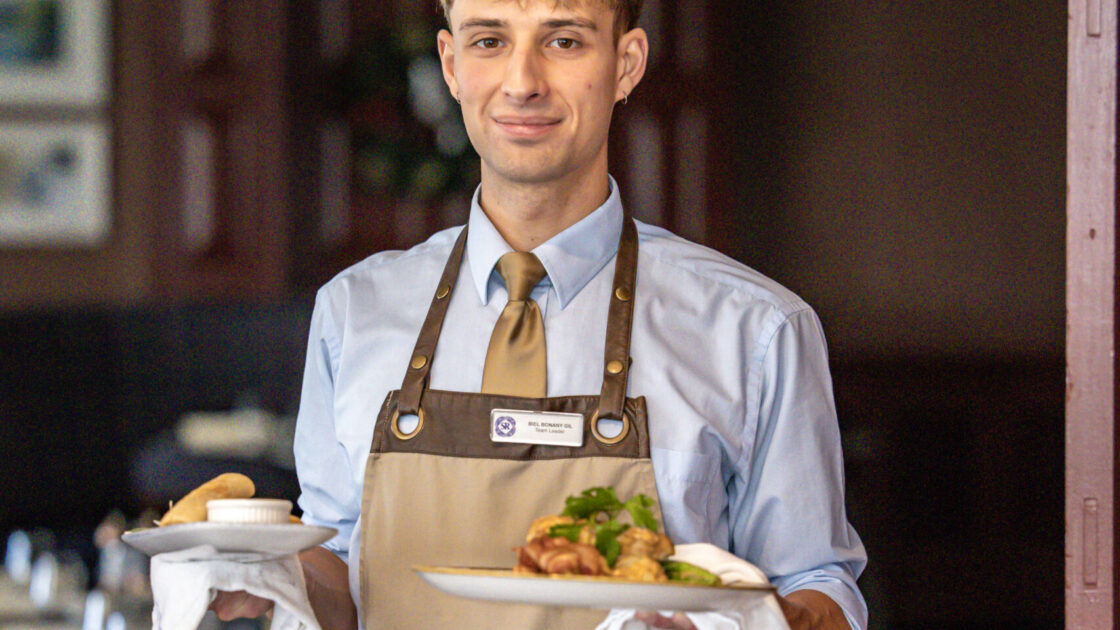 Friendly waiter in uniform serving gourmet dishes in elegant hotel dining room, offering a warm and inviting atmosphere.