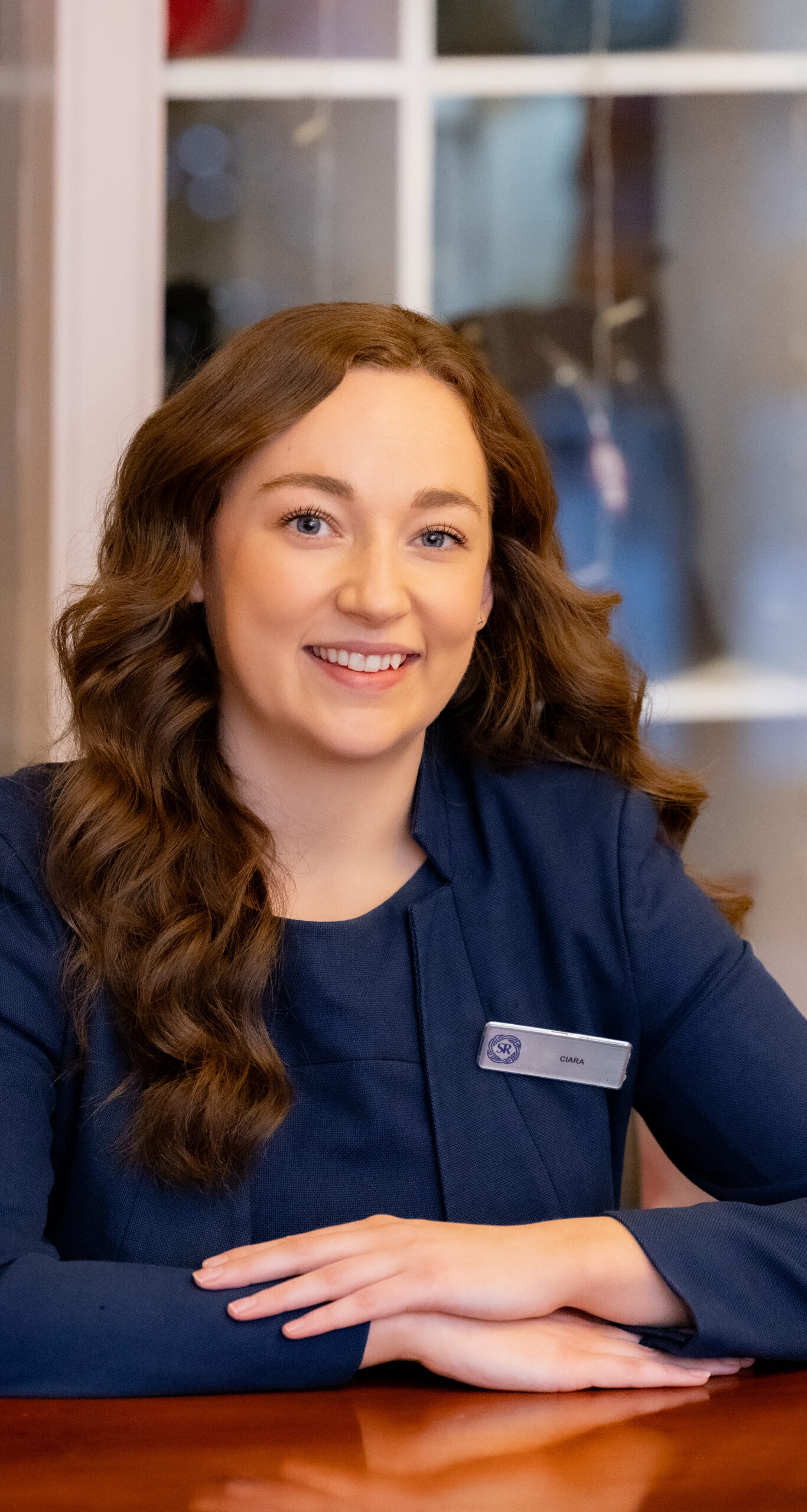 A woman with long brown hair, wearing a navy blazer and name tag, sits at a polished table and smiles at the camera. Shelves with various objects are in the background.