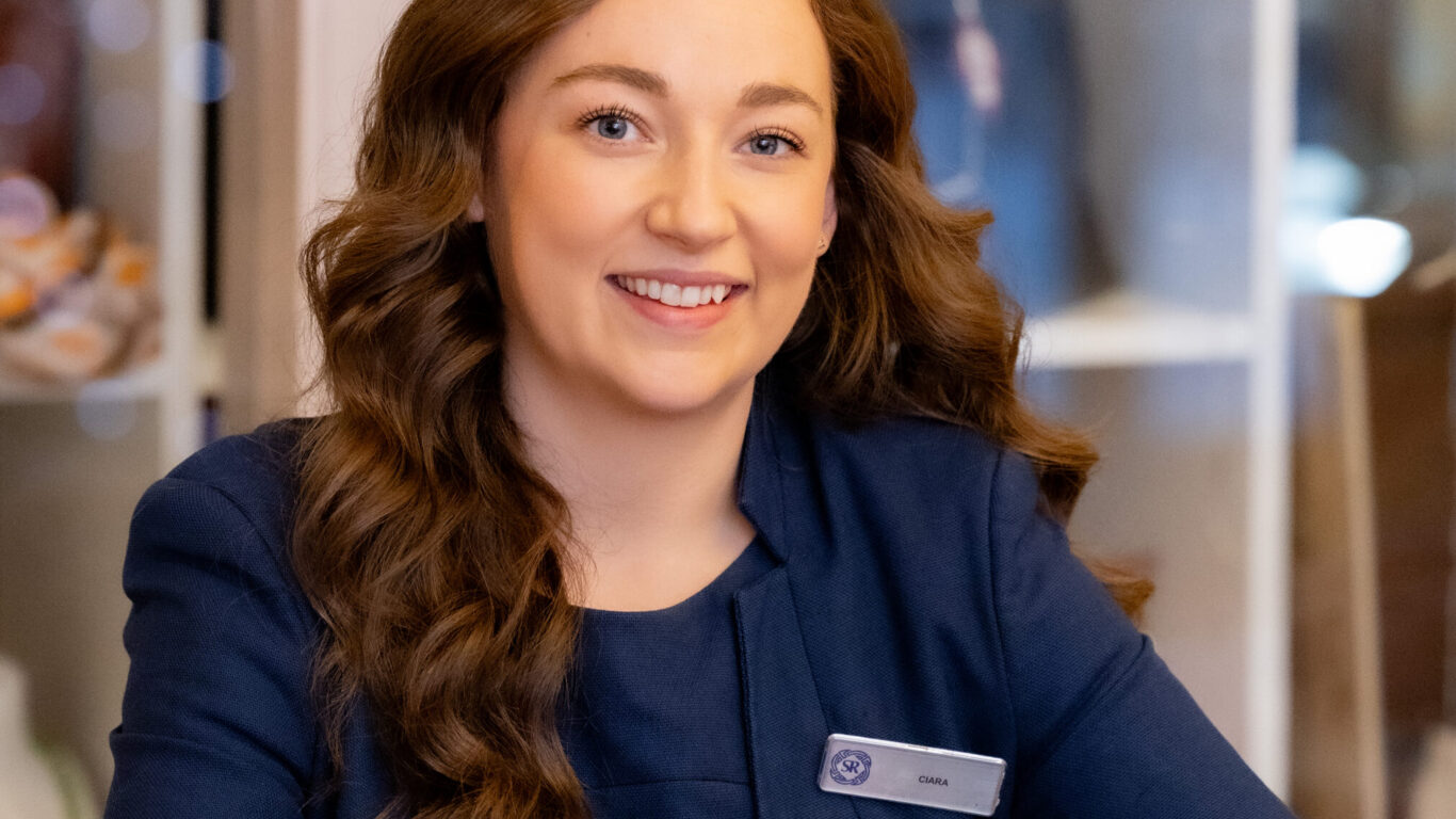 A woman with long brown hair, wearing a navy blazer and name tag, sits at a polished table and smiles at the camera. Shelves with various objects are in the background.