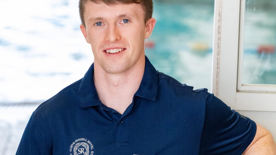 A man in a navy polo shirt with a name tag sits on a chair by an indoor swimming pool, smiling at the camera.