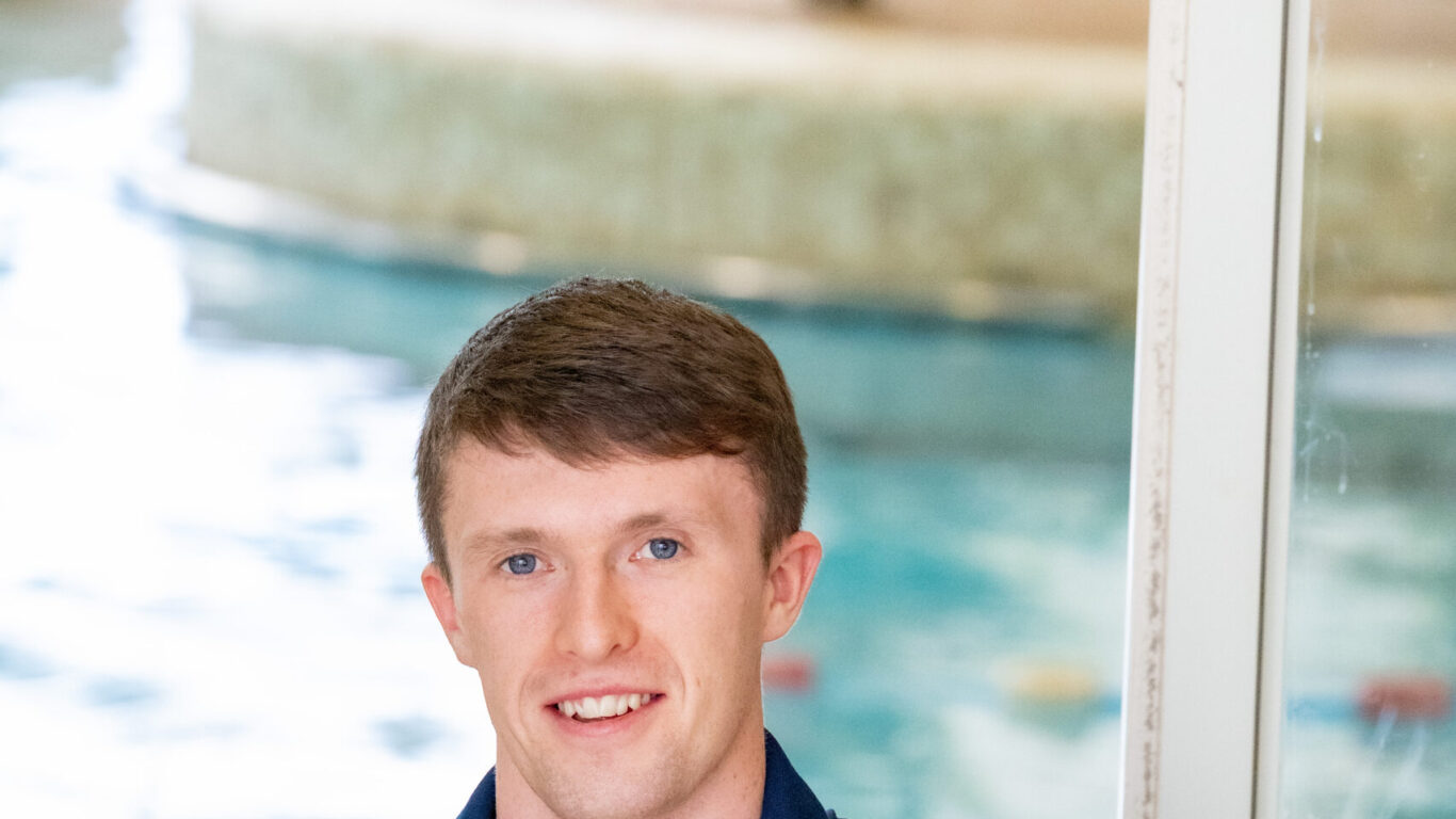A man in a navy polo shirt with a name tag sits on a chair by an indoor swimming pool, smiling at the camera.