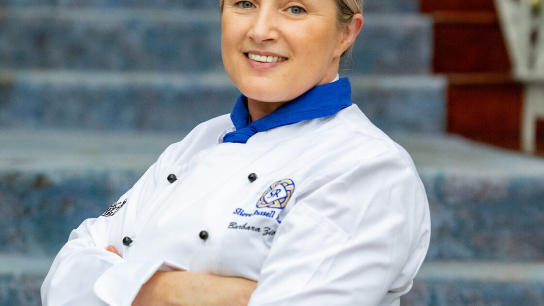 A chef in a white uniform and hat stands with folded arms, smiling, in front of a blue and red staircase.