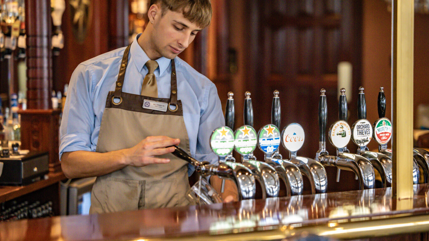 A bartender in uniform pours a drink from a beer tap at a bar, with several labeled beer taps visible on the counter.