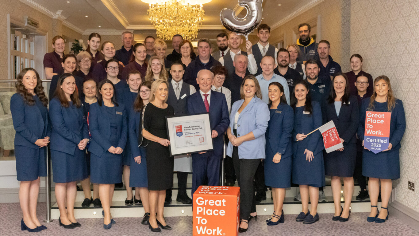A large group of professionally dressed people pose indoors, holding 