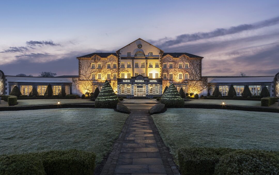 A grand hotel building with illuminated windows and a central entrance, surrounded by neatly trimmed bushes and a wide lawn at dusk.