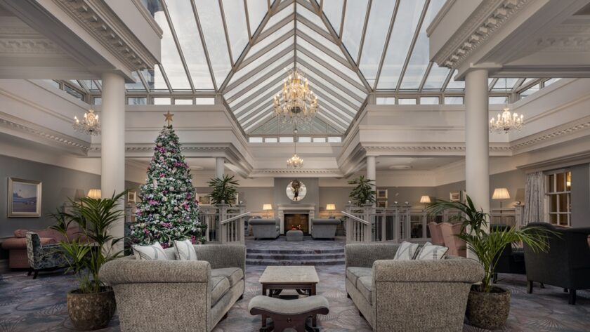Spacious hotel lobby with high glass ceiling, chandeliers, a decorated Christmas tree, sofas, armchairs, plants, and a fireplace at the far end.