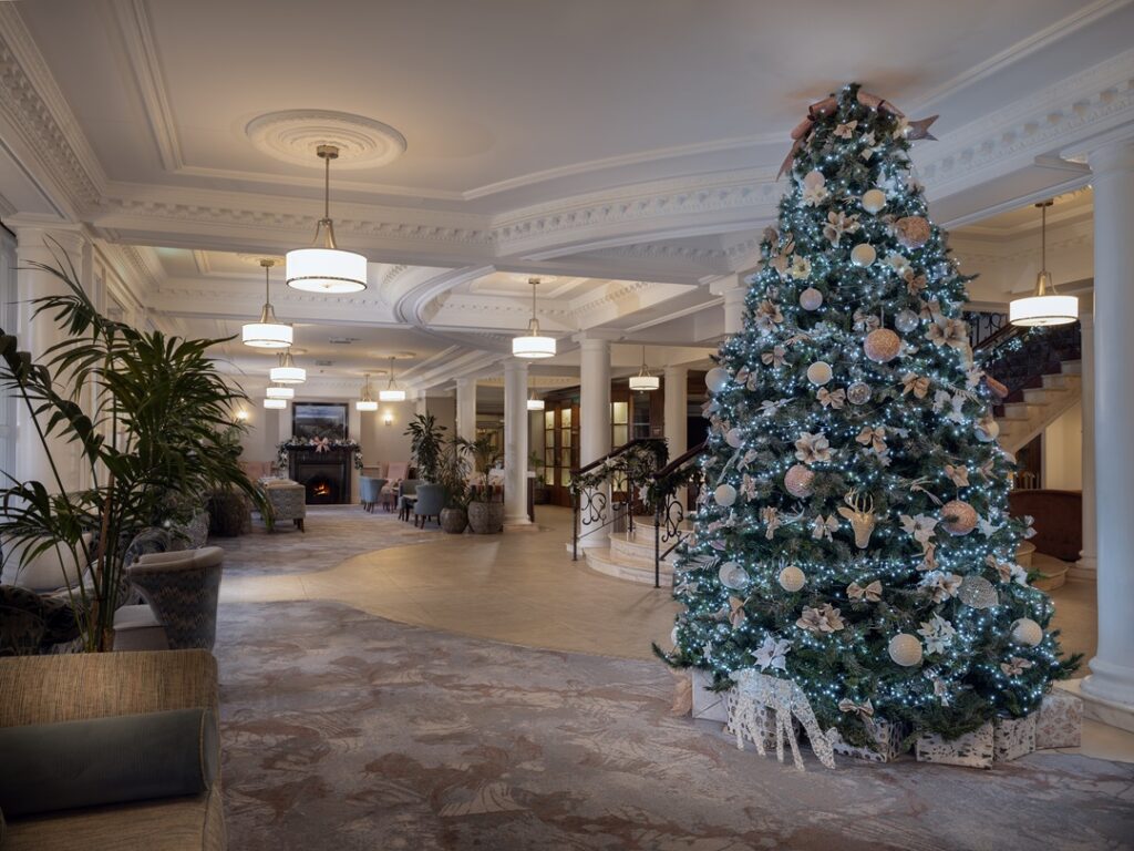 A large decorated Christmas tree stands in a spacious, elegant hotel lobby with columns, seating areas, and warm lighting.