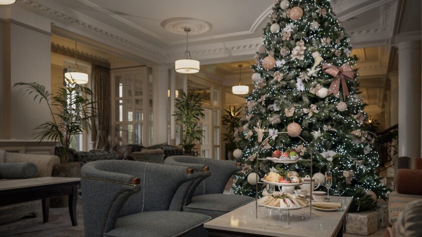 A decorated Christmas tree stands in a hotel lounge next to a table set with afternoon tea and two gray armchairs.